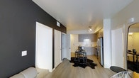 This 2-bedroom, 2-bathroom single-floor residence offers a thoughtfully designed open-concept layout that seamlessly connects the living and kitchen areas. The interior features warm-toned hardwood-style flooring that runs throughout, complementing the clean white ceilings and creating a cohesive, modern aesthetic.

The kitchen is well-appointed with grey, two-toned cabinetry, a classic white subway tile backsplash, and stainless steel appliances including a refrigerator and range. Ample counter space and a functional layout make the kitchen both practical and visually appealing. Track lighting above provides focused illumination for the cooking area.

A bold dark accent wall adds a striking contrast to the otherwise light and neutral color palette of the living space, lending the unit a contemporary edge. Multiple doors along the hallway provide access to the two bedrooms and bathrooms, offering a clear and organized flow throughout the unit. The open living area accommodates a dining or multipurpose table setup, adding to the unit's functional versatility. Overall, the space exudes a clean, modern ambiance with tasteful design choices throughout.
