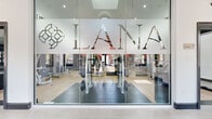 The Lana fitness center is a thoughtfully designed, single-floor facility that combines contemporary aesthetics with a full suite of workout amenities. Entry is made through a striking glass-panel facade branded with the Lana logo, offering a transparent view into the space and setting an inviting, modern tone from the outset.

Inside, the gym is anchored by dramatically vaulted ceilings that lend an open, airy quality to the interior. Decorative wooden dowels cascade from the ceiling line, creating a distinctive visual feature that adds warmth against the bold charcoal and terracotta accent walls. Light wood-look flooring runs throughout the main workout area, complemented by rubber mat zones that delineate dedicated exercise spaces.

The facility is comprehensively equipped with treadmills, ellipticals, rowing machines, cable machines, and a full rack of free weights. Ceiling fans and mounted flat-screen televisions add comfort and entertainment. Floor-to-ceiling mirrors along one wall enhance the sense of space while supporting form monitoring during workouts. A medicine ball station and yoga mat area accommodate functional and flexibility training.

A bright, tiled entryway corridor with black trim detailing and natural window views connects the main gym area, providing a clean transition into the workout space. The overall ambiance is energetic yet polished.