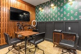 This single-room podcast studio is a thoughtfully designed recording space that balances professional functionality with distinctive interior styling. The room's most immediately recognizable design element is the deep forest-green wallpaper adorned with a repeating owl motif, which covers the upper portion of the feature wall above black wainscoting panels. This is contrasted by warm vertical wood slat paneling on the opposing wall, where a large wall-mounted flat-screen display is positioned for monitoring or content playback.

The studio is equipped for multi-participant recording, featuring a central wooden work table surrounded by black upholstered rolling chairs and multiple professional-grade boom microphone arms with headphones at each position. An audio mixing console sits atop a slatted wood credenza along one wall, providing accessible production controls. Brass wall sconces provide warm, ambient lighting throughout the room, complementing the moody, intimate atmosphere of the space.

A patterned yellow-toned carpet grounds the room, and a black-framed door with frosted glass panels provides the entry point. Large floor-to-ceiling windows along one wall allow views to an outdoor courtyard, with roller shades available for light control during recording sessions. A ring light on a stand offers additional video lighting capability.