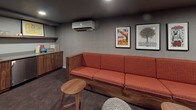 This single-floor entertainment lounge is thoughtfully designed for relaxed gatherings and media experiences. The room is anchored by a generous L-shaped sectional sofa upholstered in a warm terracotta fabric, set on a solid walnut base, and arranged to face a large wall-mounted flat-screen television. Wall-mounted speakers flank the display, ensuring an immersive audio-visual setup. Recessed ceiling lighting and a dark ceiling create an intimate, theater-like ambiance throughout the space.

The flooring features deep charcoal carpet, adding to the cozy atmosphere, while the walls are finished in a subtle textured gray that complements the overall palette. A collection of framed vintage-style posters adds personality and a retro aesthetic to the seating area. Round walnut side tables and knit pouf ottomans are thoughtfully scattered throughout for added functionality and comfort.

On the opposite end of the room, a built-in wet bar area offers walnut cabinetry, open wood shelving stocked with glassware, a sink with a modern faucet, and a compact refrigerator — ideal for entertaining. A wall-mounted smart control panel provides convenient management of the room's systems. A mini-split air conditioning unit ensures year-round comfort. The space also includes a low walnut credenza with vinyl record storage, enhancing its curated, mid-century modern character.