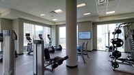 This fitness center is designed to offer a functional and comfortable workout environment. The space is anchored by a dropped ceiling with recessed panel lighting that provides bright, even illumination throughout. Structural columns are integrated into the layout, adding an architectural element while dividing the floor into distinct workout zones. Large floor-to-ceiling windows line the perimeter, flooding the space with natural light and offering expansive views of the surrounding cityscape, creating an energizing atmosphere for exercise. The flooring is a durable, dark-toned material suited for high-traffic gym use. Cardio equipment, including ellipticals and stationary machines, is positioned near the windows to take advantage of the views. Strength and resistance equipment, including benches and free-weight storage, occupies additional areas of the floor. A wall-mounted flat-screen television is also present, providing entertainment during workouts. An exercise ball and other fitness accessories are visible, indicating a versatile space for varied training routines. The overall design balances practicality with a modern, clean aesthetic.