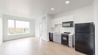 Situated in the heart of Columbus, Ohio, this efficiently designed studio at 195 E Main St offers a smart, modern layout that maximizes every square foot. The open-concept living and kitchen area is bathed in natural light courtesy of large windows that frame views of the surrounding urban streetscape, creating a bright and airy atmosphere throughout.

The kitchen is thoughtfully appointed with white upper cabinetry, gray lower cabinets, and light-toned countertops that contrast cleanly against a suite of black appliances, including a refrigerator, electric range, over-the-range microwave, and dishwasher. A stainless steel sink completes the functional workspace, making the kitchen both practical and visually cohesive.

Light wood-tone flooring runs seamlessly throughout the space, complementing the crisp white walls and lending warmth to the otherwise minimalist aesthetic. Recessed lighting adds a polished touch overhead, while clean white baseboards provide a sharp, finished look to each room.

The bathroom features a sliding door entry and includes built-in cabinetry with a glass shelf, offering tidy storage solutions. A door off the main living area provides access to additional building common spaces. The overall design is clean, contemporary, and well-suited for urban living in downtown Columbus.