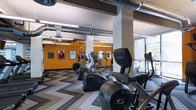 This fitness center offers a dynamic and motivating environment designed for comprehensive workouts. The space features an impressive lineup of cardio equipment, including Cybex arc trainers and elliptical machines, arranged across a stylish chevron-patterned floor that adds a modern, energetic feel underfoot. Soaring ceilings with exposed industrial ductwork and structural columns lend the space an open, airy character, while large windows along one side flood the area with natural light and provide views of the surrounding greenery. A bold orange accent wall serves as a striking visual focal point, adorned with framed sports jerseys and memorabilia that celebrate athletic achievement and inspire those working out. Ceiling fans are positioned throughout to ensure comfortable airflow during intense training sessions. The thoughtful layout provides ample space between machines, allowing for ease of movement and an uncluttered workout experience. The combination of industrial design elements, vibrant color choices, and carefully curated sports décor creates an environment that is both functionally efficient and visually engaging, making it a standout amenity for fitness enthusiasts.