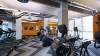 This fitness center offers a dynamic and motivating environment designed for comprehensive workouts. The space features an impressive lineup of cardio equipment, including Cybex arc trainers and elliptical machines, arranged across a stylish chevron-patterned floor that adds a modern, energetic feel underfoot. Soaring ceilings with exposed industrial ductwork and structural columns lend the space an open, airy character, while large windows along one side flood the area with natural light and provide views of the surrounding greenery. A bold orange accent wall serves as a striking visual focal point, adorned with framed sports jerseys and memorabilia that celebrate athletic achievement and inspire those working out. Ceiling fans are positioned throughout to ensure comfortable airflow during intense training sessions. The thoughtful layout provides ample space between machines, allowing for ease of movement and an uncluttered workout experience. The combination of industrial design elements, vibrant color choices, and carefully curated sports décor creates an environment that is both functionally efficient and visually engaging, making it a standout amenity for fitness enthusiasts.