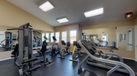 This fitness center is a thoughtfully arranged exercise space designed to support a comprehensive workout routine. The room features a dark, durable rubber floor that provides cushioning and traction throughout the entire area, creating a functional foundation suited for high-traffic use. Overhead, recessed flat-panel fluorescent fixtures illuminate the space evenly, ensuring clear visibility across all equipment zones.

The cardio section is well-stocked, featuring multiple treadmills from brands including Star Trac and Life Fitness, alongside upright and recumbent stationary bikes. A multi-station cable weight machine by Precor anchors the strength training area, accompanied by a dedicated bench for additional exercises. Full-length mirrors line one wall, visually expanding the space and allowing users to monitor their form during workouts.

Natural light filters in through vertical-blind-covered windows along one wall, adding a welcoming quality to the otherwise utilitarian space. Motivational fitness artwork is displayed on the warm beige walls, contributing to an encouraging atmosphere. A glass-door entry connects to a tiled vestibule, providing a clear transition point. The overall layout is organized and efficient, making effective use of the available floor space for a variety of fitness activities.