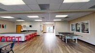 This single-floor amenity facility is thoughtfully designed to serve as a dynamic hub for recreation and socializing. The open main area is anchored by warm wood-look flooring and a drop tile ceiling with recessed panel lighting that ensures consistent, bright illumination throughout. A full-size ping pong table takes center stage, complemented by a dining area with black metal chairs and tables, ideal for group gatherings. A sleek, L-shaped wet bar with dark countertops, a double sink, and ample cabinetry lines one wall, accented by bold red metal barstools and decorative hexagonal mirrors that add a modern touch. A seating nook near the entrance features vibrant green accent chairs and views to an outdoor deck through glass double doors, flooding the corner with natural light. A dedicated lounge area offers a teal sofa, a round white coffee table, and a wall-mounted flat-screen TV, perfect for relaxing. An adjoining fitness room is visible through a glass partition and houses cardio equipment for residents' use. Multiple windows throughout the space invite natural light and offer views of the surrounding greenery, contributing to an open and welcoming atmosphere suited for both casual use and organized events.