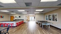 This single-floor amenity facility is thoughtfully designed to serve as a dynamic hub for recreation and socializing. The open main area is anchored by warm wood-look flooring and a drop tile ceiling with recessed panel lighting that ensures consistent, bright illumination throughout. A full-size ping pong table takes center stage, complemented by a dining area with black metal chairs and tables, ideal for group gatherings. A sleek, L-shaped wet bar with dark countertops, a double sink, and ample cabinetry lines one wall, accented by bold red metal barstools and decorative hexagonal mirrors that add a modern touch. A seating nook near the entrance features vibrant green accent chairs and views to an outdoor deck through glass double doors, flooding the corner with natural light. A dedicated lounge area offers a teal sofa, a round white coffee table, and a wall-mounted flat-screen TV, perfect for relaxing. An adjoining fitness room is visible through a glass partition and houses cardio equipment for residents' use. Multiple windows throughout the space invite natural light and offer views of the surrounding greenery, contributing to an open and welcoming atmosphere suited for both casual use and organized events.