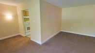 This single-floor space features a straightforward, functional layout with a living area that flows toward a partially open kitchen. The living area is finished with wall-to-wall carpet in a neutral taupe tone, providing a soft underfoot feel and a cohesive look throughout. Walls are painted in a warm off-white or light beige, contributing to a clean, understated ambiance that allows for flexible decorating.

A partial dividing wall separates the living space from the kitchen, creating a subtle sense of definition between the two areas while maintaining an open feel. The kitchen is visible through an open passageway and features white appliances, including a freestanding range, along with light wood-toned cabinetry positioned above the counters. White baseboards run along the perimeter of both spaces, adding a crisp contrast to the neutral walls and flooring.

The space is compact but practical, making efficient use of its layout. The absence of clutter and the neutral palette give the space a blank-canvas quality, well-suited for personalization. Overall, the unit presents a modest, move-in-ready environment.