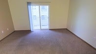 This single-floor space features a clean, functional layout centered around a generously sized living area finished with wall-to-wall carpeting in a neutral tone. The carpet provides a soft, comfortable underfoot feel and pairs well with the light yellow walls, creating a warm and understated interior palette. Smooth white baseboards frame the perimeter of the room, adding a crisp finishing detail throughout.

A prominent sliding glass door serves as the room's focal point, drawing in abundant natural light and providing direct access to a private balcony enclosed by a white railing. The balcony offers views of the surrounding building exterior, making it a practical outdoor extension of the living space. Sheer white curtains flank the sliding door, allowing diffused light to filter into the room while maintaining a sense of openness.

The walls are smooth and painted in a consistent soft yellow, contributing to a bright and airy atmosphere. The overall design is minimal and versatile, providing a neutral canvas well-suited for a variety of furnishing styles. The space presents a straightforward and practical layout with an emphasis on natural light and easy indoor-outdoor connectivity.