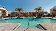 Located in Gulf Breeze, Florida, this resort-style pool area offers a stunning outdoor retreat designed for relaxation and recreation. The centerpiece is a generously sized swimming pool with brilliantly clear, turquoise water that reflects the Florida sunshine and the surrounding palm trees. Stainless steel entry rails provide easy access into the pool, while shallow wading ledges invite leisurely lounging at the water's edge.

The pool deck is finished with elegant, neutral-toned paver stones that extend throughout the surrounding area, providing both aesthetic appeal and comfortable footing. Tall, mature palm trees are strategically placed around the pool, lending a tropical ambiance and natural shade. Bright teal market umbrellas are stationed throughout the deck, accompanied by lounge chairs ideal for sunbathing.

A covered outdoor pavilion area adjacent to the pool features a ceiling fan and lush hanging basket plants, offering a shaded respite from the sun. Glass doors connect this covered space to the pool deck, creating a seamless indoor-outdoor flow. The entire pool area is enclosed by sleek black iron fencing, ensuring both safety and an open, airy feel. The multi-story residential buildings visible in the background complete the community setting of this exceptional amenity space.