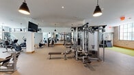 This expansive fitness center offers a comprehensive, well-appointed workout facility designed to accommodate a wide range of exercise disciplines. The open floor plan is generously proportioned, providing ample space for members to move freely between equipment zones without congestion.

The strength training area is extensively equipped with multi-use cable machines, weight racks loaded with barbells and plate-loaded stations, a full rack of dumbbells, and adjustable benches — all set against floor-to-ceiling mirrors that enhance the sense of space. A dedicated functional training zone features turf flooring and houses kettlebells, medicine balls, and other free-motion equipment, accented by an exposed brick wall that adds a rugged, industrial character.

The cardio zone lines a wall of large, grid-paned windows that flood the space with natural light and offer views of the surrounding urban streetscape. A row of Life Fitness treadmills and upright cycles make up this area. A separate room with hardwood flooring houses ICG cycling bikes and a heavy punching bag, ideal for high-intensity sessions.

Overhead, sleek black pendant lights complement the modern, neutral-toned interior. A curated gallery of black-and-white sports photography adorns one wall, adding a motivational and design-forward touch to the overall ambiance.