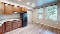 Located at 3040 Bellevue Way NE in Bellevue, Washington, this efficiently designed single-floor unit makes smart use of its compact footprint. The open-plan living, dining, and kitchen area forms the heart of the space, with light hardwood-style flooring running seamlessly throughout, lending a clean and cohesive aesthetic. Recessed lighting and a central ceiling fixture keep the area bright, while two windows along one wall invite natural light and views of surrounding greenery, creating a refreshing connection to the outdoors.

The kitchen is well-equipped with warm-toned wood cabinetry — both upper and lower — offering ample storage. A stainless steel sink with an L-shaped countertop, a built-in cooktop, and a full-size black refrigerator provide all essential cooking amenities. The layout allows the kitchen to flow naturally into the living and dining zone, making the space feel open and functional.

A dedicated bathroom includes a pedestal sink, toilet, and a separate shower/tub area, finished with neutral gray flooring and clean white walls. A hallway connects the main living area to the bathroom, with recessed lighting ensuring the corridor remains well-lit. The entry door features a solid wood finish, adding a warm, welcoming touch to this practical and neatly appointed unit.