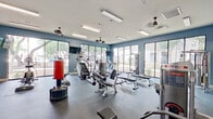 Located at 8823 Dugas Rd in San Antonio, TX, this fitness center is a single-floor facility designed to support a comprehensive workout experience. The space is finished with bold slate-blue walls and durable dark rubber flooring, creating a focused and energetic atmosphere throughout.

Large floor-to-ceiling windows line multiple walls, flooding the interior with abundant natural light and offering open views of the surrounding landscaped grounds and mature trees. Ceiling fans are distributed across the space to maintain air circulation, complementing the overhead fluorescent lighting.

The cardio zone is well-appointed with Matrix stair climbers, treadmills, ellipticals, and stationary bikes, all positioned to take advantage of the exterior views. The strength training area features a multi-station cable machine, a full rack of dumbbells neatly organized along a wall-length mirror, weight benches, and a variety of selectorized machines targeting multiple muscle groups. A freestanding boxing bag rounds out the functional training options.

Mounted flat-screen TVs are positioned throughout the space for entertainment during workouts. French doors on one side provide access to an adjacent outdoor area, while the overall layout ensures ample open floor space for movement and stretching. The fitness center offers a modern, fully outfitted environment suited for a wide range of training styles.