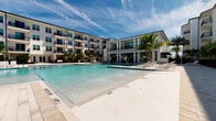 Located at 3017 Clementine Ct in Sarasota, Florida, this resort-style amenity space delivers a sophisticated outdoor living experience designed for relaxation and entertainment. The centerpiece is a generously sized zero-entry pool with fountain features, framed by swaying palm trees and the community's modern multi-story architecture, creating a vibrant Florida backdrop.

The covered outdoor lounge area is thoughtfully organized into multiple distinct zones. A seating area anchored by a marble-surround linear fireplace serves as an elegant focal point, with wicker sofas and accent pillows arranged for comfortable gathering. Mounted flat-screen televisions are positioned throughout, ensuring unobstructed viewing from multiple vantage points.

A dedicated games zone features a ping-pong table alongside high-top seating, providing a lively recreational option. Additional seating clusters with wicker armchairs and teal accent ottomans offer flexible social spaces, while dining-height tables and chairs accommodate casual meals or socializing.

The covered pavilion is finished with light blue tongue-and-groove ceilings, recessed lighting, and statement ceiling fans that provide comfortable airflow. Limestone tile flooring extends throughout, lending a clean, upscale aesthetic. Floor-to-ceiling glass walls on one side connect the outdoor space to an interior amenity area, maximizing natural light and visual continuity. The railing-lined perimeter provides open views of the pool and landscaped grounds, reinforcing the space's resort-inspired ambiance.