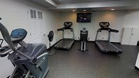 This fitness room offers a practical and functional space for cardiovascular exercise. The room is outfitted with multiple treadmills positioned side by side, a stationary recumbent bike, and an elliptical machine, providing a solid range of cardio equipment for residents or guests. A wall-mounted flat-screen television is installed at eye level, allowing users to stay entertained or informed during their workouts. A standing floor fan provides additional air circulation throughout the space. The flooring consists of durable, speckled rubber gym matting, well-suited for a high-traffic fitness environment and easy to maintain. Recessed lighting in the ceiling provides adequate illumination throughout the room. The neutral-toned walls and clean, minimal design keep the focus on functionality. A paneled door provides access to the room, and the overall layout is efficiently organized to maximize the use of the available space. This fitness room serves as a convenient, no-frills exercise facility designed to support everyday cardio training needs.
