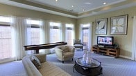 This well-appointed single-floor space in Amarillo, Texas, offers a thoughtfully designed layout that balances comfort and functionality. The main living area immediately draws attention with its elegant tray ceiling adorned with recessed lighting, adding a sense of architectural depth and refinement to the room. Warm, neutral wall tones create a welcoming ambiance that complements the plush carpeted flooring throughout. An abundance of natural light pours in through multiple large windows dressed with layered Roman shades and sheer white curtains, brightening the space and providing a light, airy feel. The living area is generously furnished with a comfortable sectional sofa, a round glass-top coffee table accented with decorative chess pieces, and a pair of accent chairs, creating an inviting conversational arrangement. A flat-screen television mounted on a media console anchors one wall, while a sleek shuffleboard table adds a playful entertainment element along another. A framed piece of wall art contributes a sophisticated finishing touch. The overall ambiance is one of relaxed elegance, making this space well-suited for both quiet evenings and social gatherings.