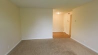 This single-floor space presents a clean, functional layout with a clear distinction between living and transitional areas. The main living area is finished with wall-to-wall carpet in a neutral beige tone, providing a comfortable and quiet underfoot feel. Crisp white baseboards line the perimeter, complementing the soft, off-white walls that lend a bright and open atmosphere throughout.

A transitional hallway extends from the main living space, finished with hardwood-style flooring that offers a practical contrast to the carpeted area. The hallway is illuminated by a flush-mount ceiling light, ensuring adequate visibility. A louvered bifold door is visible along the hall, likely concealing a closet or utility space, and a wall-mounted panel — possibly an intercom or thermostat — is positioned nearby.

The entry door is visible at the end of the hallway, framed cleanly within the neutral color scheme. The overall ambiance of the space is understated and move-in ready, with a simple, well-maintained interior that provides a neutral canvas adaptable to a variety of furnishing styles and uses.
