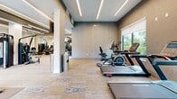 Located at 7430 Lyons Road in Wellington, Florida, this well-appointed fitness center offers a comprehensive and modern workout environment designed to meet a wide range of exercise needs. The space is thoughtfully laid out across a single floor, providing ample room for both cardiovascular training and strength conditioning without feeling crowded.

The interior design is characterized by a contemporary aesthetic, featuring a dark tray ceiling with striking linear LED lighting that runs in parallel strips, creating a clean and energizing ambiance. Patterned carpet tiles in neutral tones cover the floor, adding texture while maintaining a polished appearance. Structural columns are integrated into the layout and are complemented by large mirrors that visually expand the space and allow for form-checking during workouts.

The cardio zone is equipped with treadmills, elliptical machines, and stationary cycling bikes, all positioned near floor-to-ceiling windows that frame lush, tropical outdoor views — a hallmark of the South Florida setting. The strength training area features a robust selection of Hoist selectorized machines and cable systems, along with a free weights section complete with a dumbbell rack, adjustable bench, and stability ball storage.

Additional amenities include wall-mounted flat-screen televisions, a water fountain station accessible through glass-panel doors, and dedicated rubber flooring mats in the free weights area. Natural light streams in generously through multiple large windows, enhancing the open and inviting atmosphere of this well-equipped facility.