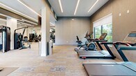 Located at 7430 Lyons Road in Wellington, Florida, this well-appointed fitness center offers a comprehensive and modern workout environment designed to meet a wide range of exercise needs. The space is thoughtfully laid out across a single floor, providing ample room for both cardiovascular training and strength conditioning without feeling crowded.

The interior design is characterized by a contemporary aesthetic, featuring a dark tray ceiling with striking linear LED lighting that runs in parallel strips, creating a clean and energizing ambiance. Patterned carpet tiles in neutral tones cover the floor, adding texture while maintaining a polished appearance. Structural columns are integrated into the layout and are complemented by large mirrors that visually expand the space and allow for form-checking during workouts.

The cardio zone is equipped with treadmills, elliptical machines, and stationary cycling bikes, all positioned near floor-to-ceiling windows that frame lush, tropical outdoor views — a hallmark of the South Florida setting. The strength training area features a robust selection of Hoist selectorized machines and cable systems, along with a free weights section complete with a dumbbell rack, adjustable bench, and stability ball storage.

Additional amenities include wall-mounted flat-screen televisions, a water fountain station accessible through glass-panel doors, and dedicated rubber flooring mats in the free weights area. Natural light streams in generously through multiple large windows, enhancing the open and inviting atmosphere of this well-equipped facility.