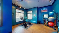 Located at 1 Springtime Blvd SW in Huntsville, Alabama, this single-floor fitness studio delivers an energizing workout environment defined by its bold teal and cobalt blue walls, warm hardwood flooring, and dynamic layout designed for active use.

The main workout area is bright and open, drawing in natural light through multiple large windows fitted with horizontal blinds. Track lighting overhead illuminates the space efficiently. The room is equipped with stationary cycling bikes featuring integrated digital screens, positioned on protective floor mats for focused cardio sessions. A dedicated equipment wall showcases a well-organized rack stocked with VTX medicine balls in varying sizes, a full set of color-coded resistance bands, and stability balls — providing versatile options for strength and functional training.

Colorful abstract artwork adorns the walls, adding an uplifting, creative energy to the space. A corner area features a floor lamp and additional cycling equipment, offering a more intimate training zone with outdoor views through surrounding windows.

Adjacent to the main floor is a welcoming reception or lounge area, painted in a matching teal palette. This area features a coffee station, hand sanitizer dispenser, towel service, and a striking neon sign reading "Eyes on the Finish Line," reinforcing the motivational atmosphere throughout the studio.