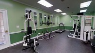 This fitness room offers a practical and functional space designed to support a variety of workout routines. The room features sage green walls accented with white crown molding, giving the space a clean and refreshing aesthetic. Durable rubber flooring covers the entire area, providing a safe and resilient surface ideal for exercise equipment and high-impact activity. Overhead, recessed rectangular panel lights ensure the room is well-lit throughout. A multi-station strength training machine anchors one side of the room, offering users a range of resistance exercises in a compact footprint. A pull-up and dip station stands nearby, providing additional bodyweight training options. A treadmill is positioned toward the back of the room, offering dedicated space for cardiovascular workouts. A weight bench with additional equipment rounds out the selection on the opposite side. A series of wall-mounted mirrors line one wall, allowing users to monitor their form during exercise while also contributing to a sense of openness in the room. A small informational sign is mounted on the wall, indicating guidelines or instructions for use. Overall, the space is thoughtfully arranged to maximize functionality and accommodate a diverse range of fitness activities.