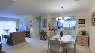 This single-floor residence offers a thoughtfully designed open-plan layout that seamlessly connects the living, dining, and kitchen areas, creating a spacious and cohesive environment. The interior is dressed in soft, neutral tones — warm beige walls and plush carpet flooring — lending the space a calm and inviting ambiance throughout.

The dining area serves as a central focal point, anchored by a white oval dining table surrounded by slatted wooden chairs with upholstered seats. Above, an elegant chandelier with frosted glass shades adds a refined touch. A decorative curio cabinet nearby adds character and charm to the space.

The kitchen is bright and functional, benefiting from generous natural light streaming through curtained windows. It features light wood cabinetry, modern appliances including a built-in microwave, and ample counter space, making it well-suited for everyday cooking and entertaining alike.

The adjacent living area provides a comfortable retreat, complete with a sofa, television, and a delicate lamp that contributes to the warm, homely atmosphere. Wall-hung artwork and mirrors add personality and depth to the interiors.

A door visible from the main living space hints at additional rooms beyond, while the overall layout ensures a smooth, connected flow between all areas of this well-appointed single-level home.