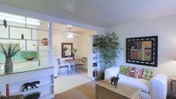 This single-floor space showcases an open-concept design that seamlessly connects the living and dining areas, creating a cohesive and functional layout. The living area features neutral-toned carpeting and is furnished with a white upholstered sofa accented by decorative pillows, a woven rattan coffee table, and a tall indoor bamboo plant that adds a natural, organic element to the space. A framed piece of wall art provides a focal point on one wall, while built-in white shelving units flank the open passage to the dining area, offering both display and storage opportunities.

The dining area is accessed through a wide, open doorway framed by white trim, giving the space an airy, connected feel. A ceiling fan with a light fixture illuminates the dining space, which features a round table paired with wrought iron chairs. A decorative mirror adorns the wall, enhancing the sense of depth. The light-colored flooring in the dining area contrasts gently with the carpeted living space. Overall, the interior presents a calm, well-organized ambiance with thoughtful decorative touches throughout.