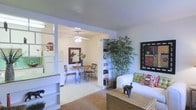 This single-floor space showcases an open-concept design that seamlessly connects the living and dining areas, creating a cohesive and functional layout. The living area features neutral-toned carpeting and is furnished with a white upholstered sofa accented by decorative pillows, a woven rattan coffee table, and a tall indoor bamboo plant that adds a natural, organic element to the space. A framed piece of wall art provides a focal point on one wall, while built-in white shelving units flank the open passage to the dining area, offering both display and storage opportunities.

The dining area is accessed through a wide, open doorway framed by white trim, giving the space an airy, connected feel. A ceiling fan with a light fixture illuminates the dining space, which features a round table paired with wrought iron chairs. A decorative mirror adorns the wall, enhancing the sense of depth. The light-colored flooring in the dining area contrasts gently with the carpeted living space. Overall, the interior presents a calm, well-organized ambiance with thoughtful decorative touches throughout.