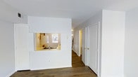 This two-floor space is defined by a clean, minimalist aesthetic anchored by crisp white walls and rich, dark hardwood-style flooring that runs throughout the main level. A standout architectural detail is the pass-through window cut into the wall between the living area and the kitchen, creating an open sightline that connects the two spaces while maintaining defined zones. The kitchen, visible through the pass-through, features a sink positioned beneath a window that allows natural light to filter in, contributing to the overall brightness of the space. White cabinetry and appliances complement the kitchen's light and airy feel. The hallway extending from the main living area leads to multiple rooms, each accessed through white paneled doors with gold-toned hardware, adding a subtle warmth to the otherwise neutral palette. The layout appears well-organized, with thoughtful separation between living, kitchen, and additional rooms. The staircase visible in the background indicates seamless access to the second floor. Overall, the space presents a functional and tidy environment with consistent design choices that give it a cohesive and welcoming character.