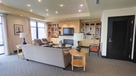 This thoughtfully designed single-floor space presents a sophisticated and inviting lounge environment with a cohesive contemporary aesthetic. At the heart of the room, a tiled fireplace serves as a focal point, flanked by a wall-mounted flat-screen television and framed by warm-toned built-in shelving units that provide both display and storage space. The built-ins are finished in a light natural wood, lending the space a clean and modern character while offering a sense of structure and organization.

The seating arrangement is generously appointed, featuring a large upholstered sofa paired with matching armchairs in a neutral taupe palette, complemented by accent chairs in a striking burnt orange that add visual interest and contrast. Rounded wood coffee and side tables anchor the seating area, adding warmth and functionality. Elegant table lamps provide ambient lighting, supplemented by recessed ceiling lights that illuminate the tray ceiling detail above.

Large windows along one wall allow natural light to filter into the space, enhancing its open and airy atmosphere. The carpeted flooring in a subtle pattern adds texture underfoot while contributing to the room's overall sense of comfort. The overall ambiance is polished yet approachable, making the space well-suited for relaxed gatherings or quiet leisure.