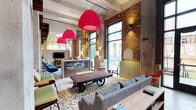 This striking single-floor space masterfully blends industrial and contemporary design elements to create a warm, energetic environment. Exposed brick walls and raw concrete ceilings establish an authentic industrial character, while oversized floor-to-ceiling windows flood the interior with natural light and offer views of the surrounding streetscape. Eye-catching crimson pendant lights hang dramatically from the ceiling, serving as bold focal points that inject vibrant color into the space. The lounge area is anchored by a distinctive reclaimed wood coffee table mounted on industrial cart wheels, paired with plush gray sofas and eclectic accent chairs in teal and green hues. A patterned area rug defines the seating zone, adding texture and visual interest underfoot. Floor lamps with unique architectural forms are strategically placed throughout, contributing both warm ambient lighting and sculptural appeal. A reception or service counter with a display case sits at the heart of the space, suggesting a hospitality or co-working function. The open floor plan allows for a natural flow between seating areas, while the layered lighting — combining pendant, floor, and wall-mounted fixtures — creates a welcoming ambiance suitable for both social interaction and relaxed conversation.