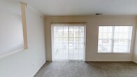 This single-floor space offers a clean, functional layout designed to maximize comfort and natural light. The main living area is finished with wall-to-wall carpet in a neutral tone, creating a warm and inviting atmosphere throughout. A prominent sliding glass door serves as a focal point, fitted with vertical blinds that allow for flexible light control while providing direct access to the outdoors. Adjacent to the sliding door, a large picture window further enhances the sense of openness, flooding the room with daylight and offering views of the surrounding greenery. The walls are painted in a soft, neutral beige that complements the light carpeting and contributes to an airy, understated aesthetic. Crown molding runs along the upper perimeter of the living area, adding a refined architectural detail to the otherwise straightforward design. Ceiling-mounted HVAC vents are integrated discreetly, ensuring year-round climate comfort. The overall layout is open and versatile, lending itself well to a variety of furniture arrangements and uses. The abundance of natural light, neutral finishes, and outdoor connectivity make this a practical and pleasant living space.