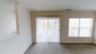 This single-floor space offers a clean, functional layout designed to maximize comfort and natural light. The main living area is finished with wall-to-wall carpet in a neutral tone, creating a warm and inviting atmosphere throughout. A prominent sliding glass door serves as a focal point, fitted with vertical blinds that allow for flexible light control while providing direct access to the outdoors. Adjacent to the sliding door, a large picture window further enhances the sense of openness, flooding the room with daylight and offering views of the surrounding greenery. The walls are painted in a soft, neutral beige that complements the light carpeting and contributes to an airy, understated aesthetic. Crown molding runs along the upper perimeter of the living area, adding a refined architectural detail to the otherwise straightforward design. Ceiling-mounted HVAC vents are integrated discreetly, ensuring year-round climate comfort. The overall layout is open and versatile, lending itself well to a variety of furniture arrangements and uses. The abundance of natural light, neutral finishes, and outdoor connectivity make this a practical and pleasant living space.