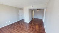 This single-story unit is finished with dark hardwood-style flooring that runs consistently throughout all living areas, creating a cohesive and grounded aesthetic. The walls are painted in a neutral gray tone, complemented by crisp white baseboards and trim, giving the interior a clean, contemporary feel. Textured popcorn ceilings are present throughout.

The open living areas are connected seamlessly, with ceiling fans providing both lighting and air circulation across multiple rooms. A dedicated living space features a large sliding glass door dressed with vertical blinds, allowing natural light to filter in while offering outdoor access. Additional rooms benefit from windows fitted with blinds, maintaining a consistent look throughout.

The galley-style kitchen is equipped with black appliances, including a refrigerator and electric range, paired with warm-toned laminate countertops and white cabinetry. A small window above the counter brings in natural light.

The bedroom areas are generously proportioned, with at least one room featuring a walk-in closet. The bathroom includes a tiled tub and shower combination, a toilet, vanity with laminate countertop, and a mirrored medicine cabinet, offering functional everyday convenience.