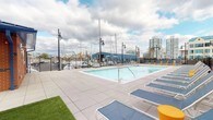 Located at 28 Southfield Ave in Stamford's waterfront South End neighborhood, this amenity level offers a thoughtfully designed collection of outdoor and transitional spaces. The centerpiece is a generous outdoor pool deck finished in large-format light gray pavers, lined with blue lounge chairs and framed by striking views of a nearby marina, sailboat masts, and the Stamford skyline — a setting that reinforces the property's waterfront character.

Adjacent to the pool area, a dedicated lawn games zone features lush artificial turf enclosed by sleek black metal fencing. Cornhole boards are set up for casual recreation, and the space is surrounded by the building's attractive red brick and white paneling exterior, with private balconies visible above.

Interior corridors connecting these outdoor spaces are finished with patterned blue-and-gray carpet, clean white baseboards, and warm neutral walls. Glass-panel doors with full-height sidelights flood the transitional entryways with natural light while offering direct sightlines to the pool deck and marina beyond. Framed artwork accents the hallways, and exit signage and fire safety equipment are cleanly integrated. The overall design balances modern residential aesthetics with practical amenity functionality, creating an inviting and cohesive outdoor living experience.