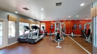 This fitness center is designed to support a comprehensive workout experience within a single-floor layout. The space is defined by striking bold red accent walls that energize the environment, complemented by a neutral-toned flooring that provides a durable, easy-to-maintain workout surface. Recessed lighting throughout the ceiling ensures the room is well-lit, while large windows along one wall allow natural light to filter in, creating an open and motivating atmosphere. Striped window valances add a decorative touch that balances the boldness of the walls. A full-wall mirror on one side visually expands the space and allows users to monitor their form during exercise. The cardio section is well-appointed with multiple treadmills and elliptical machines, offering options for various aerobic workouts. A multi-station cable and strength training machine occupies a central area of the room, providing a versatile option for full-body resistance training. The layout is thoughtfully organized to allow users to move freely between equipment. Overall, the fitness center presents a functional, energetic, and welcoming environment suited for a range of fitness routines.