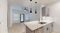 This single-floor space presents a thoughtfully designed interior with a consistent modern aesthetic carried throughout. The kitchen serves as a standout feature, outfitted with gray shaker-style cabinetry accented by matte black hardware, white quartz countertops, and a subtle tile backsplash. Stainless steel appliances — including an electric range, over-the-range microwave, dishwasher, and refrigerator — provide a polished, functional workspace. A stainless steel undermount sink with a contemporary faucet completes the well-appointed kitchen.

Light wood-tone luxury vinyl plank flooring runs throughout the living and sleeping areas, creating a warm, cohesive feel that contrasts cleanly with the bright white walls. The living area benefits from natural light through a window fitted with white blinds, while a modern ceiling fan with integrated lighting provides both airflow and illumination. Gray panel interior doors add a subtle design accent along the hallway, which connects the rooms in a straightforward layout.

The bathroom is finished in a clean, minimal style, featuring a tub-shower combination with large-format white tile surround, a single-sink vanity with a black-framed mirror, and light flooring that ties into the rest of the unit. Overall, the space presents a crisp, move-in-ready environment with consistent modern finishes.