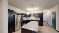 Located in Wyoming, Michigan, this single-floor unit offers a thoughtfully designed open-concept layout that seamlessly connects the kitchen and living areas. The kitchen is a standout feature, showcasing dark espresso cabinetry, granite countertops, a large center island, and a full suite of stainless steel appliances including a refrigerator, range, microwave, and dishwasher. A built-in wine rack and pendant lighting add refined detail to the space. Light wood-style flooring runs throughout the main living areas, complementing the neutral wall tones and creating a clean, contemporary aesthetic. The living area is generously proportioned, illuminated by a ceiling fan with light fixture and open sight lines to the kitchen. Multiple bedrooms feature plush carpeting and windows that allow natural light to filter in, offering views of the surrounding neighborhood. The bathroom is well-appointed with a granite-topped vanity, dark cabinetry, a full tub-shower combination, and wood-style flooring. Throughout the unit, dark door and trim accents provide a modern contrast against the light walls, contributing to a cohesive and stylish interior design. This unit combines functional layout with quality finishes for comfortable everyday living.