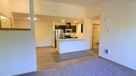 This modern single-floor residence offers a thoughtfully designed open-concept layout that seamlessly connects the kitchen and living areas. The kitchen is a standout feature, equipped with stainless steel appliances including a refrigerator, microwave, and range, complemented by sleek dark cabinetry and a light-toned countertop peninsula that doubles as a breakfast bar. Recessed lighting illuminates the kitchen, highlighting the clean, contemporary aesthetic. The transition from the kitchen flows naturally into the living area, which features warm laminate wood flooring that gives way to plush carpeting, creating distinct yet cohesive zones within the open floor plan. The neutral, light-toned walls throughout enhance the sense of brightness and openness. A glimpse into the adjoining bathroom reveals additional functional space with tiled finishes. The overall ambiance is clean, modern, and move-in ready, with a design that balances practicality and contemporary style, making it a versatile and comfortable living environment.