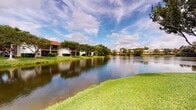 This single-floor residence is nestled within a tranquil lakeside community, offering a setting defined by natural beauty and a calming atmosphere. The exterior environment is a standout feature, with a picturesque lake at the heart of the community, its still waters reflecting the sky and the surrounding canopy of mature trees. Well-maintained green lawns extend to the water's edge, creating a park-like setting that enhances the overall sense of peaceful living. Neighboring buildings are thoughtfully integrated into the landscape, maintaining a low-rise profile that preserves open sightlines and a connection to the outdoors. The community's lush, tropical-style landscaping is consistent with a warm Florida-climate setting, providing residents with a resort-like ambiance year-round. The single-floor layout of the unit offers the convenience of easy, step-free living throughout. While interior imagery was limited, the overall character of the community suggests a space designed for comfortable, relaxed living with the ever-present backdrop of scenic waterfront views just outside.