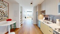 Located at 3025 John F. Kennedy Boulevard in Philadelphia's University City neighborhood, this single-floor residence showcases a thoughtfully designed contemporary interior with a clean, minimalist aesthetic. Warm hardwood flooring flows continuously throughout the space, anchoring each area with a cohesive, inviting tone.

The kitchen is efficiently laid out with light wood cabinetry extending to the ceiling, stainless steel appliances including a range and built-in microwave, a deep single-basin sink, and a dark tiled backsplash that adds subtle contrast to the otherwise neutral palette. The layout opens toward a bright corridor lined with generous built-in storage closets, providing ample organization space near the entry.

The living and sleeping areas are oriented toward floor-to-ceiling windows that frame expansive views of the Philadelphia skyline, flooding the rooms with natural light. Open shelving and a compact dining area with a round table and green stools contribute to the functional yet stylish atmosphere.

The bathroom features large-format neutral floor tiles, a tub-shower combination with full wall tile surround, a natural wood vanity with an undermount sink, and a frameless mirror with accent lighting, completing the space with a refined, spa-like simplicity.