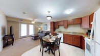 This single-floor residence showcases a thoughtfully designed open-concept layout that seamlessly integrates the kitchen, dining, and living areas into one cohesive and comfortable space. The kitchen is outfitted with rich, warm-toned wood cabinetry, granite-look countertops, a double sink, and a white dishwasher, providing both functionality and aesthetic appeal. A pendant light fixture hangs above, adding a refined touch to the cooking and prep area. Adjacent to the kitchen, the dining area accommodates a round wooden table with matching dark chairs, elegantly set for meals and centered beneath a classic pendant light. The living area flows naturally from the dining space, featuring plush carpeted flooring, a gray sofa, a coffee table, and a ceiling fan that contributes to the room's comfort. A set of French doors at the far end of the living area floods the space with natural light and provides access to the outdoors, enhancing the open and airy ambiance. The combination of tile flooring in the kitchen and dining area and carpet in the living room creates a clear yet harmonious zoning between spaces. Overall, the residence offers a warm, functional, and inviting atmosphere well-suited for everyday living.