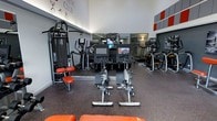 This athletic club fitness center is thoughtfully designed to support a wide range of workout routines. The space features high ceilings that create an open, energizing atmosphere, complemented by bright overhead lighting that ensures excellent visibility throughout. The flooring consists of durable rubber matting, providing both safety and comfort during exercise sessions.

The facility is outfitted with an impressive array of modern equipment. Spin bikes with integrated digital screens occupy a central position on the floor, allowing members to engage in immersive cycling experiences. Behind them, a row of treadmills lines the mirrored rear wall, which serves to visually expand the space and allow users to monitor their form. To one side, a cable strength training machine with orange upholstered seating adds a bold accent. Elliptical cross-trainers and additional cardio machines round out the equipment selection along the perimeter.

The color scheme of gray, white, and orange is carried throughout the space, from the accent wall panels to the equipment upholstery and signage, giving the facility a cohesive, professional identity. The overall layout is organized and spacious, allowing for comfortable movement between stations.