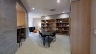 This single-floor space is thoughtfully designed to serve as both a collaborative and relaxed retreat. The main area centers around a sleek black rectangular table surrounded by modern chairs with light wooden legs, making it well-suited for group discussions or casual meetings. A large wooden shelving unit lines the back wall, featuring open cubbies filled with books, decorative objects, and artistic pieces, adding character and visual interest to the room. Recessed ceiling lighting, complemented by a floor lamp in the corner, creates a warm and focused ambiance throughout. Adjacent to the main table area is a cozy lounge nook furnished with a cushioned armchair, a small ottoman in a bold orange tone, and a side table — ideal for quieter, individual use. A bar-height counter with stools is partially visible along one wall, providing an additional area for informal work or conversation. The space features neutral gray carpeting and light-toned walls, contributing to a clean, contemporary aesthetic. The overall design balances functionality with comfort, making it a versatile environment suited for both focused work and leisurely gatherings.