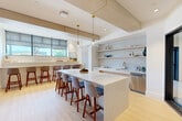 This thoughtfully designed clubroom in Beverly, MA offers a sophisticated gathering space with a seamless blend of modern aesthetics and functional amenities. The open-plan layout is anchored by light hardwood flooring and exposed wood beam accents on the ceiling, creating a warm yet contemporary atmosphere throughout.

The kitchen area is a standout feature, centered around a large white quartz island with seating for several bar-height chairs in a mid-century modern style. A textured subway tile backsplash lines the walls behind open floating shelves, which display glassware and décor. Brass-accented pendant lights hang above the island, complementing the gold-toned fixtures at the sink. A row of windows along one wall floods the space with natural light, and a built-in counter with additional bar seating runs beneath them.

Adjacent to the kitchen, the lounge and recreation area features a full-size pool table in a rich wood finish with a blue felt surface, illuminated by a striking Sputnik chandelier with brass arms. A deep blue textured accent wall anchors this zone, paired with a wall-mounted flat-screen TV, built-in shelving, and a cozy seating nook. A tufted leather banquette provides additional seating nearby, making this clubroom an ideal space for socializing and entertainment.