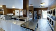 This communal laundry facility is designed with practicality and convenience in mind, offering residents everything needed for a seamless laundry experience. The space features a row of top-load washing machines neatly arranged along one wall, providing ample capacity for multiple users. A large stainless steel folding table occupies the center of the room, offering generous workspace for sorting and folding laundry. The walls are finished in a warm brown and cream color scheme, giving the space a clean and welcoming atmosphere. Flush-mounted ceiling lights provide bright, even illumination throughout. A vending machine is conveniently positioned toward the back of the room, offering easy access to laundry supplies. Informational signage and posted instructions are visible on the walls, ensuring users can easily follow facility guidelines. Light-toned floor tiles contribute to the bright and clean aesthetic of the space. A door with a glass panel at the rear allows natural light to filter in, adding to the overall openness of the room. The layout is thoughtfully organized to allow comfortable movement between machines and the folding area, making this laundry facility both efficient and user-friendly.