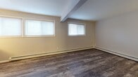 This single-floor unit offers a practical and well-maintained layout designed for comfortable everyday living. The interior is finished with wood-look plank flooring throughout, providing a warm and cohesive aesthetic across all rooms. Neutral gray and beige tones on the walls create a clean, versatile backdrop that adapts easily to various furnishing styles.

The entry foyer welcomes you through a solid wood door and features a flush-mount ceiling light and baseboard heating, setting a functional tone from the start. The main living area is generously proportioned, with two windows fitted with blinds that allow for natural light while maintaining privacy. Exposed beam-like architectural details add subtle character to the space.

A secondary room, finished with darker wood-look flooring and gray walls, connects seamlessly to other areas of the unit and offers flexible use as a bedroom or additional living space. The kitchen, visible in the floor plan, features warm wood-toned cabinetry.

The bathroom is efficiently arranged, complete with a wall-mounted sink, toilet, a shower-tub combination, and a mirrored medicine cabinet with a wood frame. Baseboard heating is present here as well, ensuring warmth throughout. Overall, this unit presents a clean, move-in-ready space with consistent finishes and practical amenities.