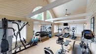 This dedicated fitness center is designed to support a comprehensive workout experience within an architecturally striking space. The room is defined by its soaring vaulted ceiling, accented by exposed white beams that lend a clean, structured aesthetic. Large arched windows positioned high along the walls flood the interior with abundant natural light, offering pleasant views of surrounding greenery and creating an open, energizing atmosphere. A ceiling fan provides comfortable air circulation throughout.

The space is thoughtfully outfitted with a wide variety of exercise equipment to accommodate diverse fitness routines. Cardio options include multiple treadmills and stationary bikes, while a rowing machine adds further variety. A well-stocked dumbbell rack, stability balls, and additional strength training equipment ensure users have everything needed for a full-body workout. Wall-mounted mirrors expand the sense of space and allow for form monitoring during exercise. A flat-screen television is mounted on the wall, offering entertainment during workouts.

The neutral-toned carpeted flooring, cream-colored wood-panel walls, and shuttered windows contribute to a warm yet functional ambiance. The combination of high architectural ceilings, ample natural light, and comprehensive equipment makes this fitness center both practical and inviting.