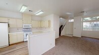 Located at 3468 Dry Gulch Dr in Laughlin, Nevada, this two-story home is designed with a clean, neutral palette throughout. The main level opens into a combined living and kitchen area, where wood-look flooring in the kitchen transitions to plush carpeting in the living space. A front entry door with a transom window allows natural light to filter into the foyer, which sits adjacent to a staircase with a white balustrade leading to the upper floor.

The kitchen is outfitted with cream-toned cabinetry offering generous upper and lower storage, white appliances including a range, built-in microwave, dishwasher, and refrigerator, along with light-toned countertops and a tile backsplash. A peninsula counter separates the kitchen from the carpeted living area, creating an open, functional layout. Flush-mounted fluorescent lighting illuminates the space overhead.

Upstairs, the bedrooms are carpeted and bright, each featuring windows with vertical blinds that invite natural light. The primary bedroom includes a ceiling fan and a spacious bathroom with a wide dual-sink vanity, a large mirror, and wood-look flooring. Additional bedrooms offer vaulted ceilings and ample wall space, making them versatile and comfortable.