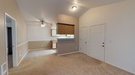 This single-floor residence offers an open and functional layout designed for comfortable everyday living. The entry foyer features tile flooring and leads directly into the main living area, which is finished with soft wall-to-wall carpeting that extends throughout the space. A defining architectural feature is the vaulted ceiling, which lends a sense of height and openness to the interior. The living area is outfitted with a ceiling fan, providing both comfort and light. A centrally located kitchen peninsula with a pass-through counter connects the kitchen to the living and dining areas, creating a cohesive open-concept flow ideal for entertaining or daily use. The kitchen is equipped with warm wood-toned upper cabinetry and light-colored lower cabinets, complemented by a countertop workspace. Neutral beige wall tones are consistent throughout, contributing to a clean, understated aesthetic. The space benefits from a straightforward, practical design that maximizes usability. Overall, the home presents a welcoming and well-organized interior with architectural details that enhance the sense of space and light.